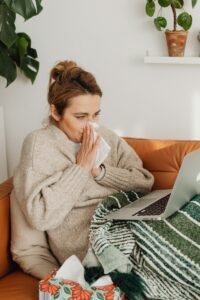 Woman in cozy sweater using laptop on sofa, surrounded by indoor plants, while blowing nose.
